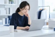 梱包用の箱がある明るい仕事場で、ノートPCを使って自社ショップの販売を確認する運営者