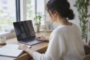 自然光の机でノートとノートパソコンを使い、サイトの記事を編集する私服の女性